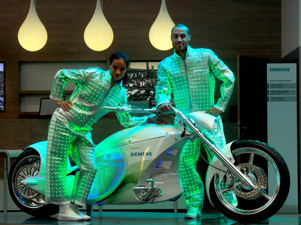 Zwei Models in von Leuchtdioden beleuchteten Anzgen posieren  auf dem Stand der Firma Siemens neben einem Elektromotorrad, dem OCC eChopper.