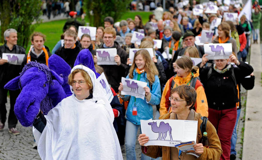 Demonstranten gehen mit einem Kamelkostm und Bildern, auf denen ein Kamel abgebildet ist, zum Haupteingang der Messe, um eine Auseinandersetzung mit der Verteilung von Vermgen in der Gesellschaft von den Kirchen zu fordern.