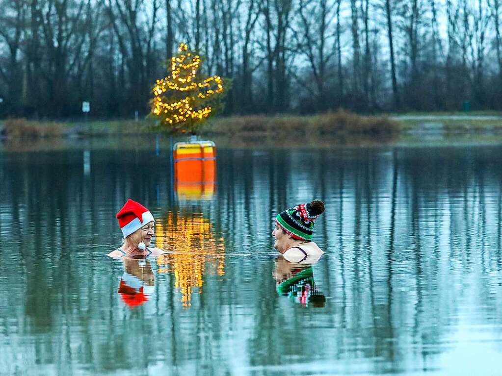 Zwei Frauen stehen kurz nach Tagesanbruch im Wasser des Schwarzachtalsee, whrend im Hintergrund ein Weihnachtsbaum auf einer Boje leuchtet.