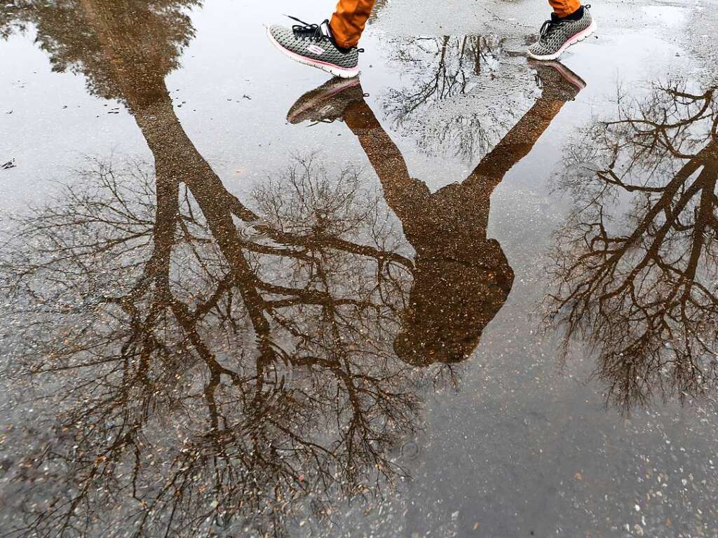 Eine Frau geht durch eine Regenpftze, in der sich kahle Bume spiegeln.