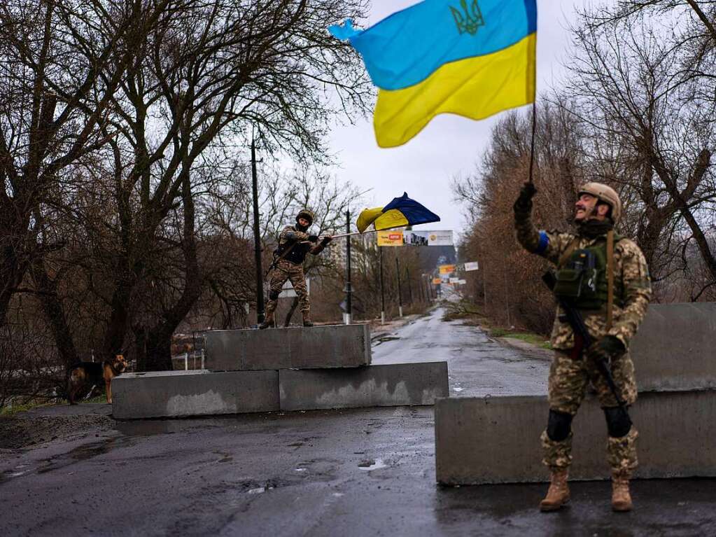 Zwei ukrainische Soldat stehen an einer Straenblockade und halten ukrainische Fahnen hoch.