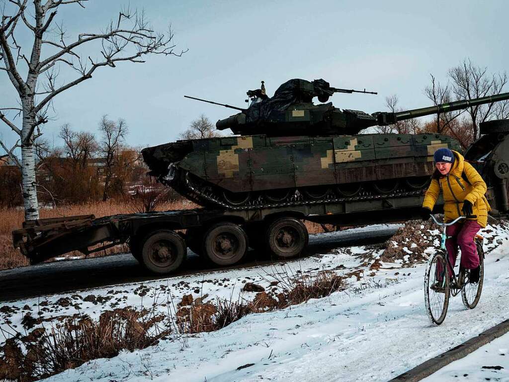 Im Winter 2023 fhrt eine Frau mit dem Fahrrad durch Kupiansk – vorbei an einem Lkw, der einen T-64-Panzer transportiert.