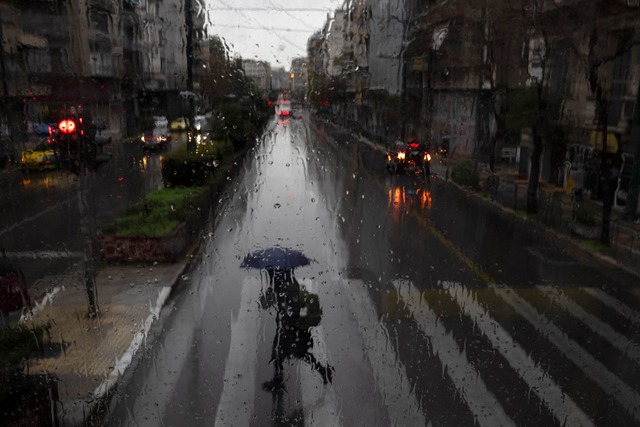 Eine Frau mit Regenschirm &uuml;berquert eine Stra&szlig;e in Athen.  | Foto: Petros Giannakouris/AP/dpa